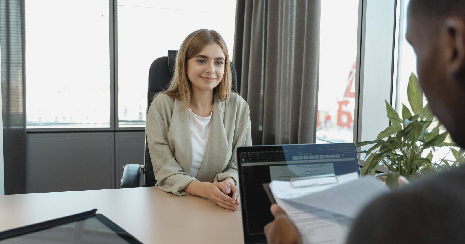Светловолосая женщина сидит напротив темнокожего мужчины, который читает её резюме и проводит собеседование.