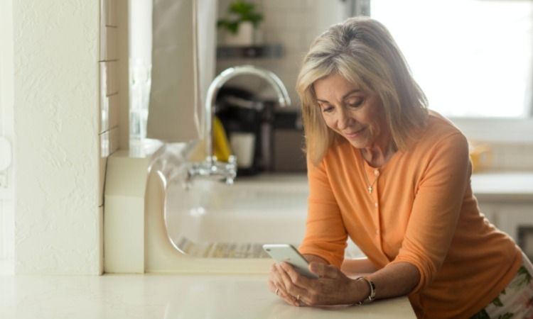 Женщина в оранжевой рубашке опирается о стол и смотрит на телефон.