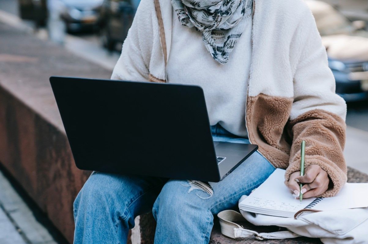 Женщина сидит на улице с ноутбуком и делает заметки о вакансиях