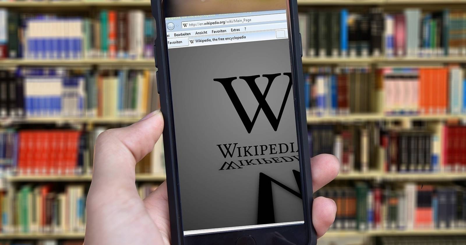 Рука держит телефон с открытой страницей Википедии на фоне книжного шкафа