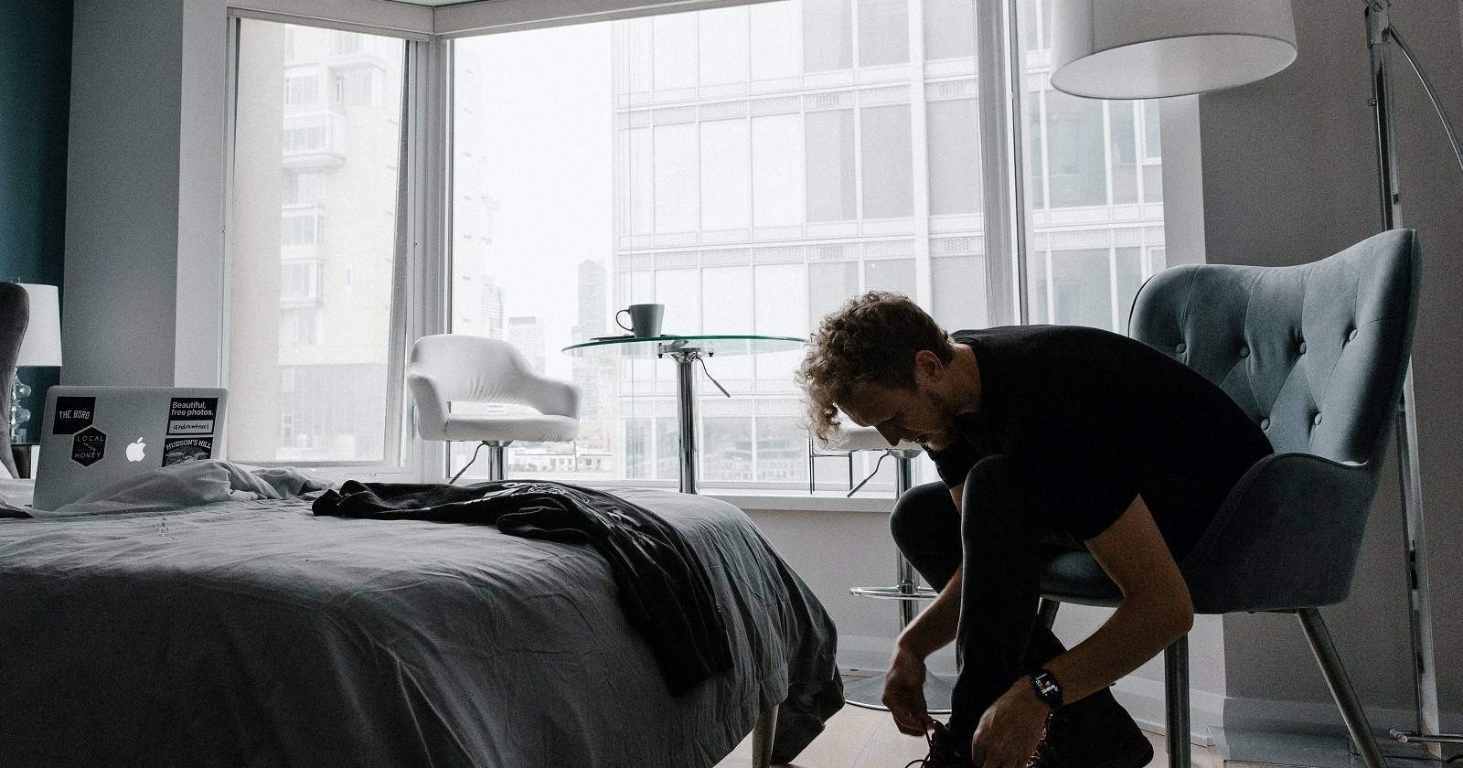 Мужчина наклоняется, чтобы завязать шнурок в хорошо освещённой комнате рядом с компьютером и чашкой кофе на фоне. 