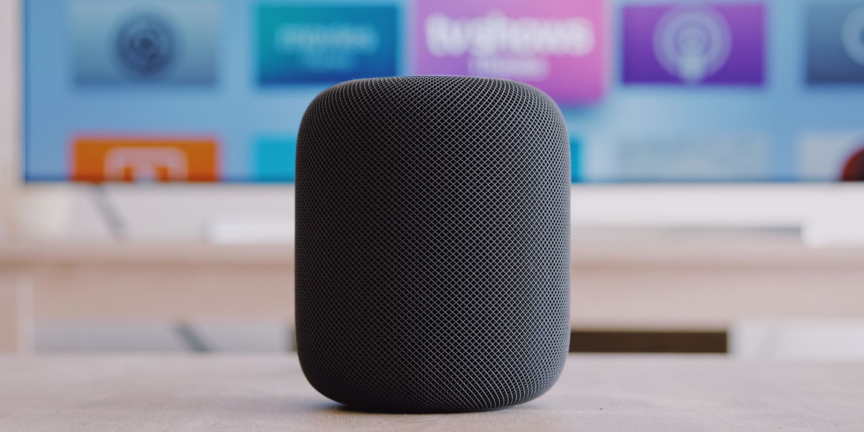 Apple HomePod тёмного цвета на столе, на фоне Apple TV