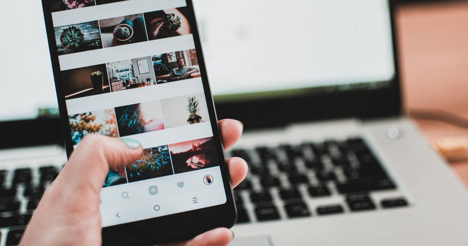 Рука держит телефон с открытым Instagram, на заднем плане ноутбук
