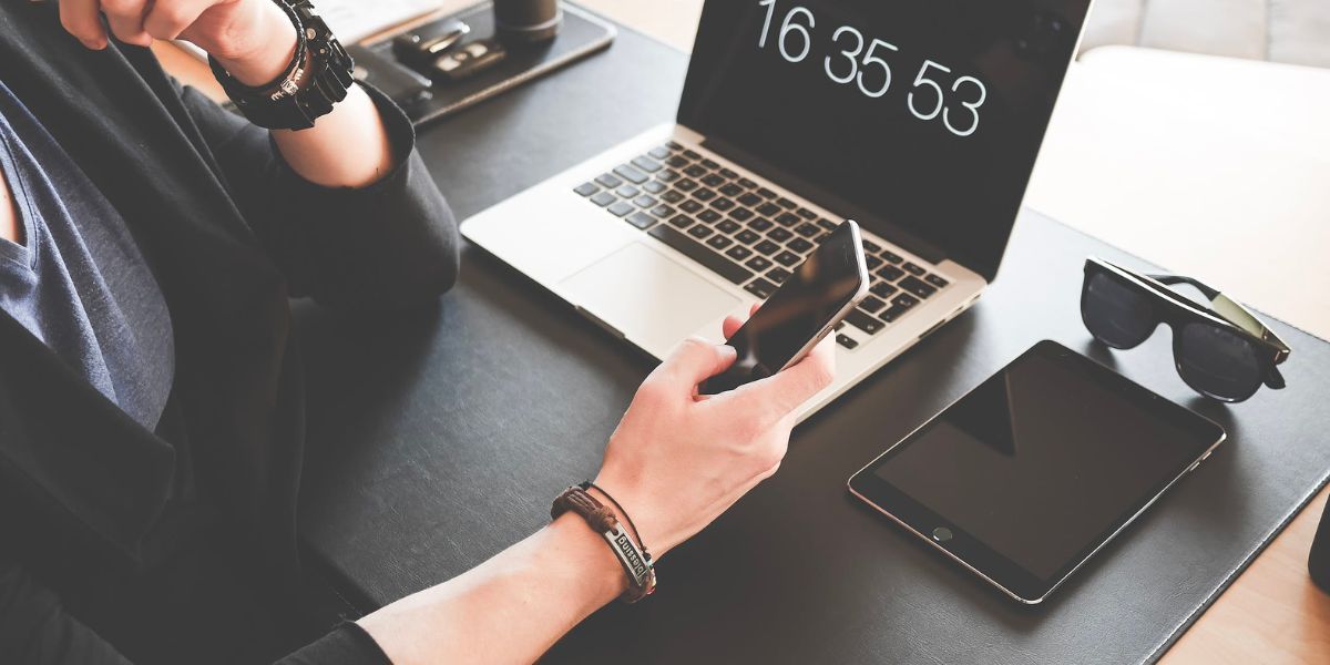 Рука держит iPhone, на столе MacBook и iPad