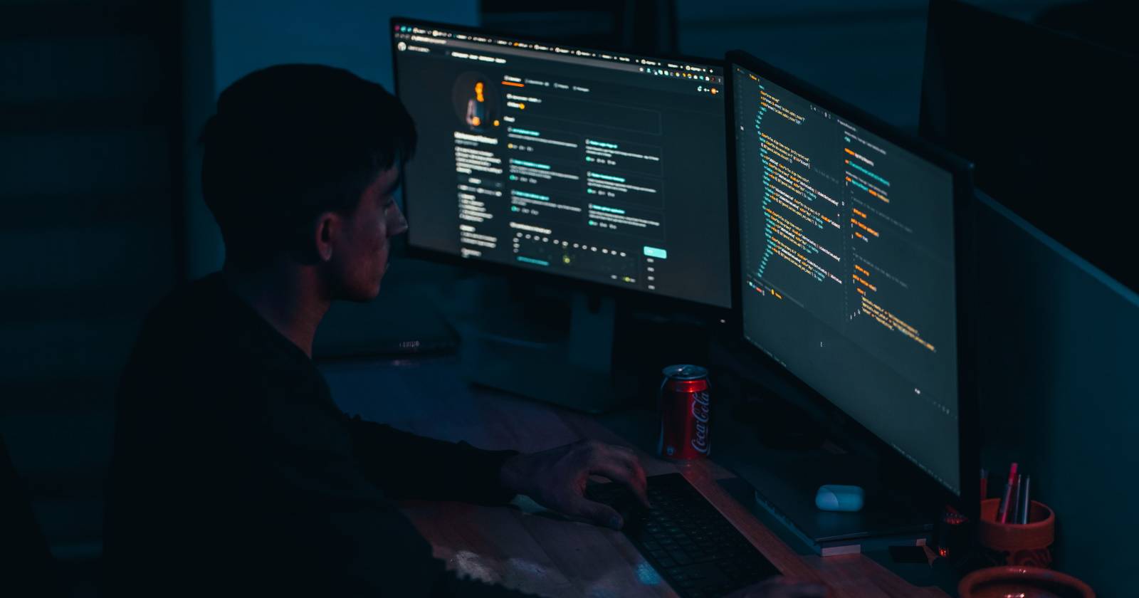 Человек пишет код в тёмной комнате перед тремя мониторами
