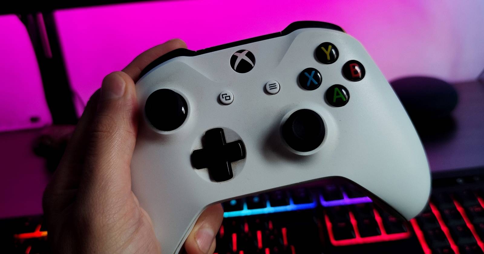 Человек держит белый беспроводной контроллер Xbox на фоне подсвеченной клавиатуры и монитора