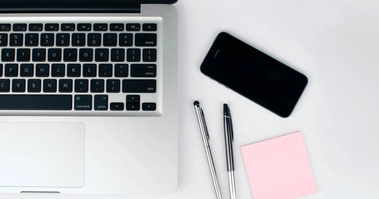 Ноутбук, смартфон, ручки и розовая стикер-заметка на белом столе