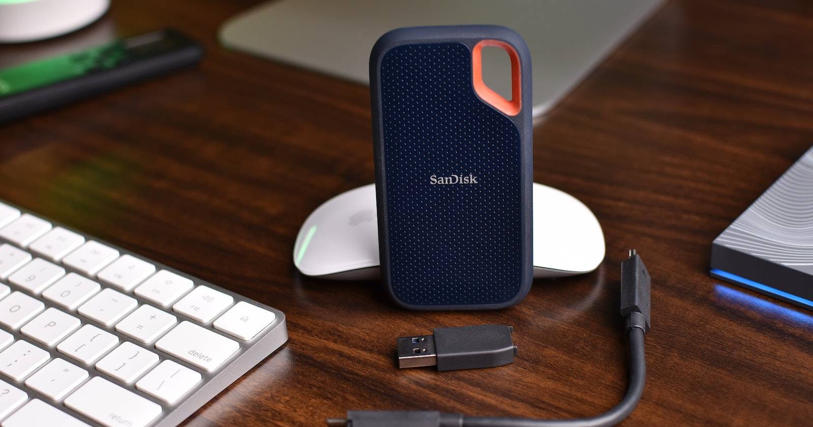 Sandisk SSD beside a USB drive, a cable, an Apple mouse, and a Magic Keyboard