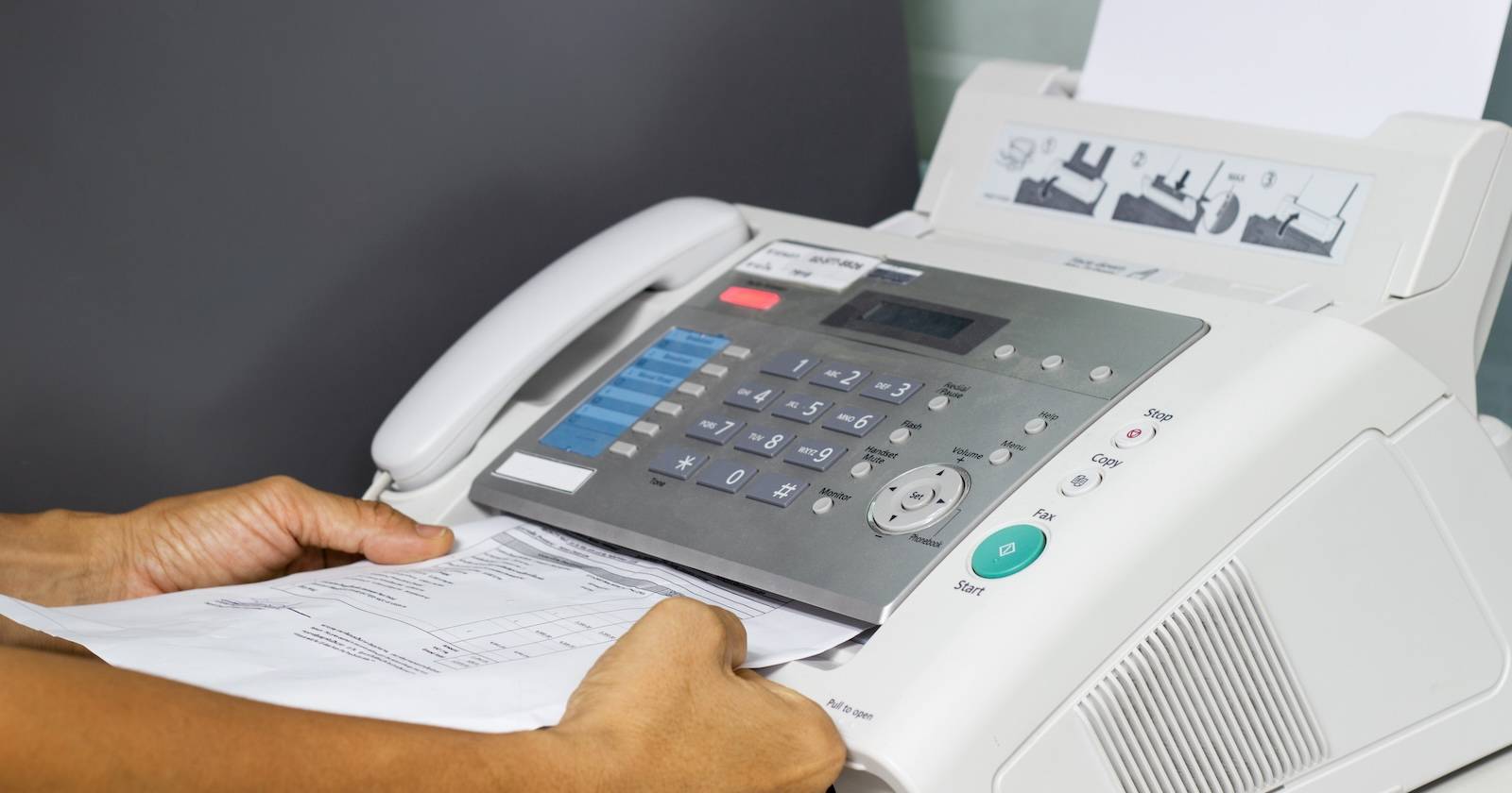 Руки вкладывают лист бумаги в громоздкий факс‑аппарат с телефонной трубкой и панелью кнопок.