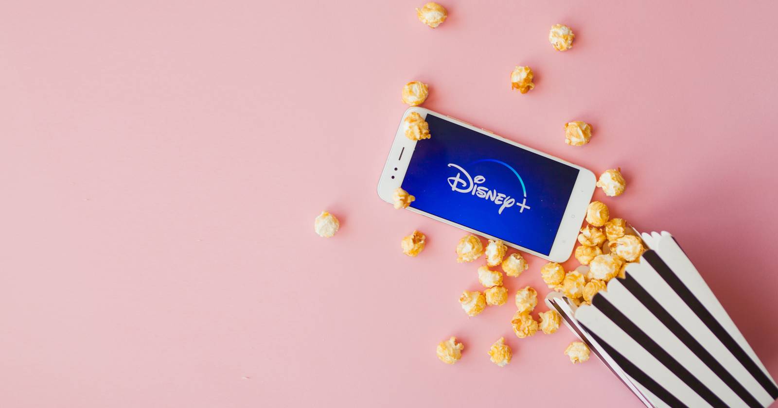 Логотип Disney+ на телефоне рядом с коробкой попкорна на розовом фоне