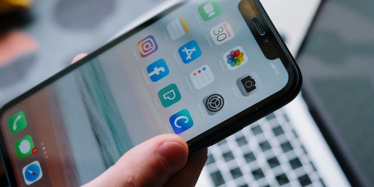 Рука держит iPhone с домашним экраном, на фоне ноутбук