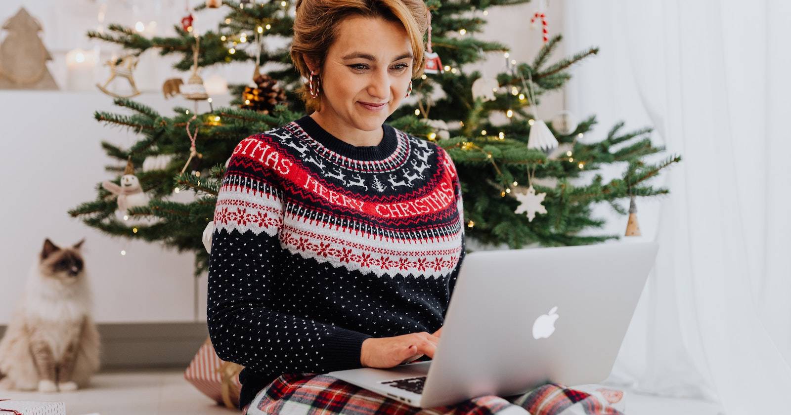 Женщина улыбается, работая на ноутбуке рядом с украшенной рождественской ёлкой