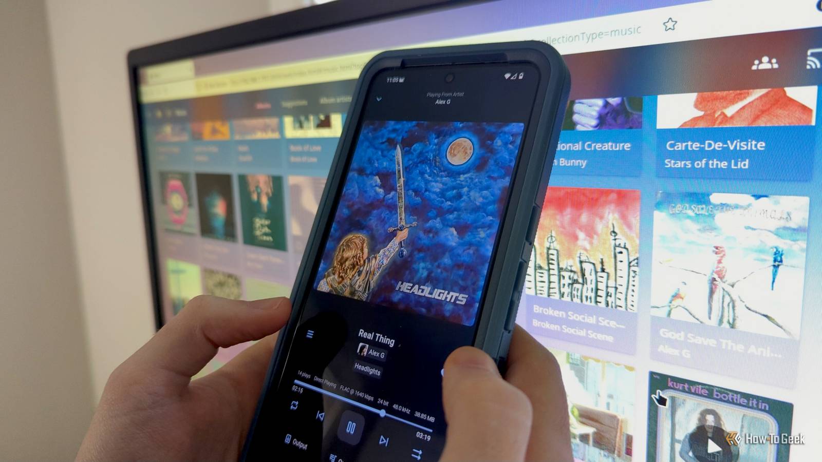 Руки человека держат Android-телефон с приложением Finamp на фоне экрана компьютера.