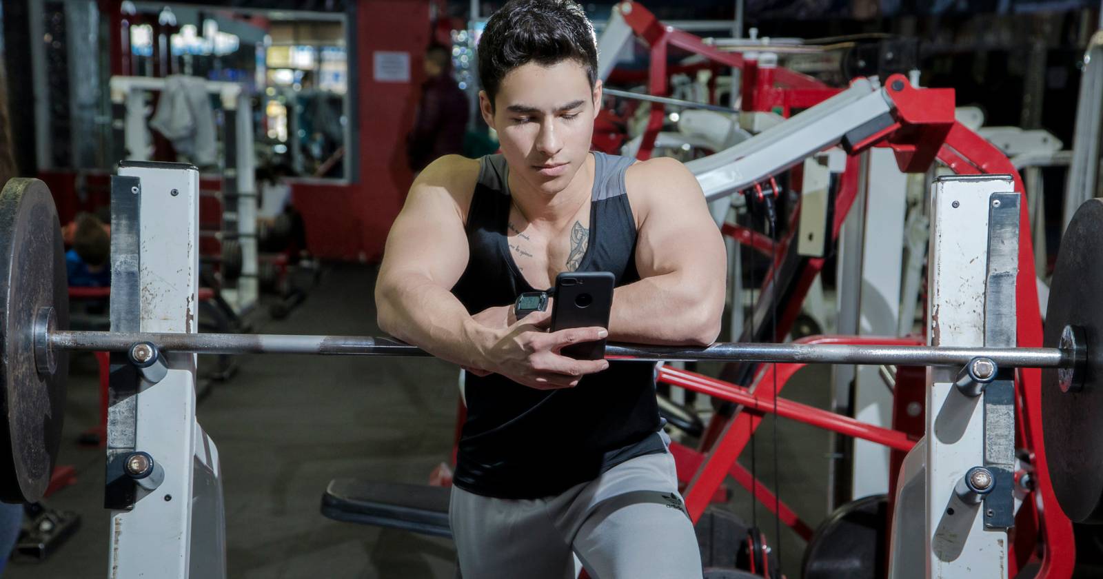 Мужчина в спортзале пользуется смартфоном, отдыхая на штанге