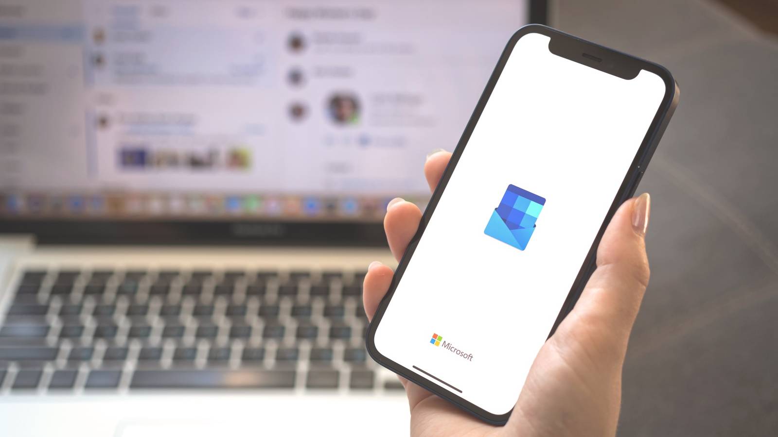 Мобильный телефон с логотипом приложения Outlook, удерживаемый перед ноутбуком.