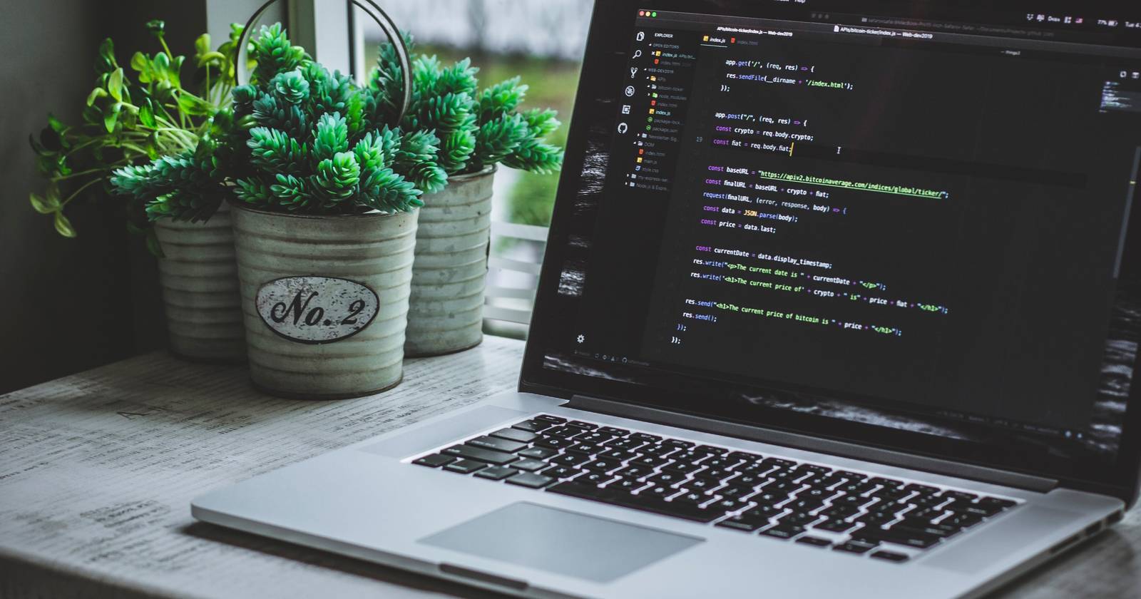 Ноутбук на столе у окна с JavaScript‑кодом на экране.