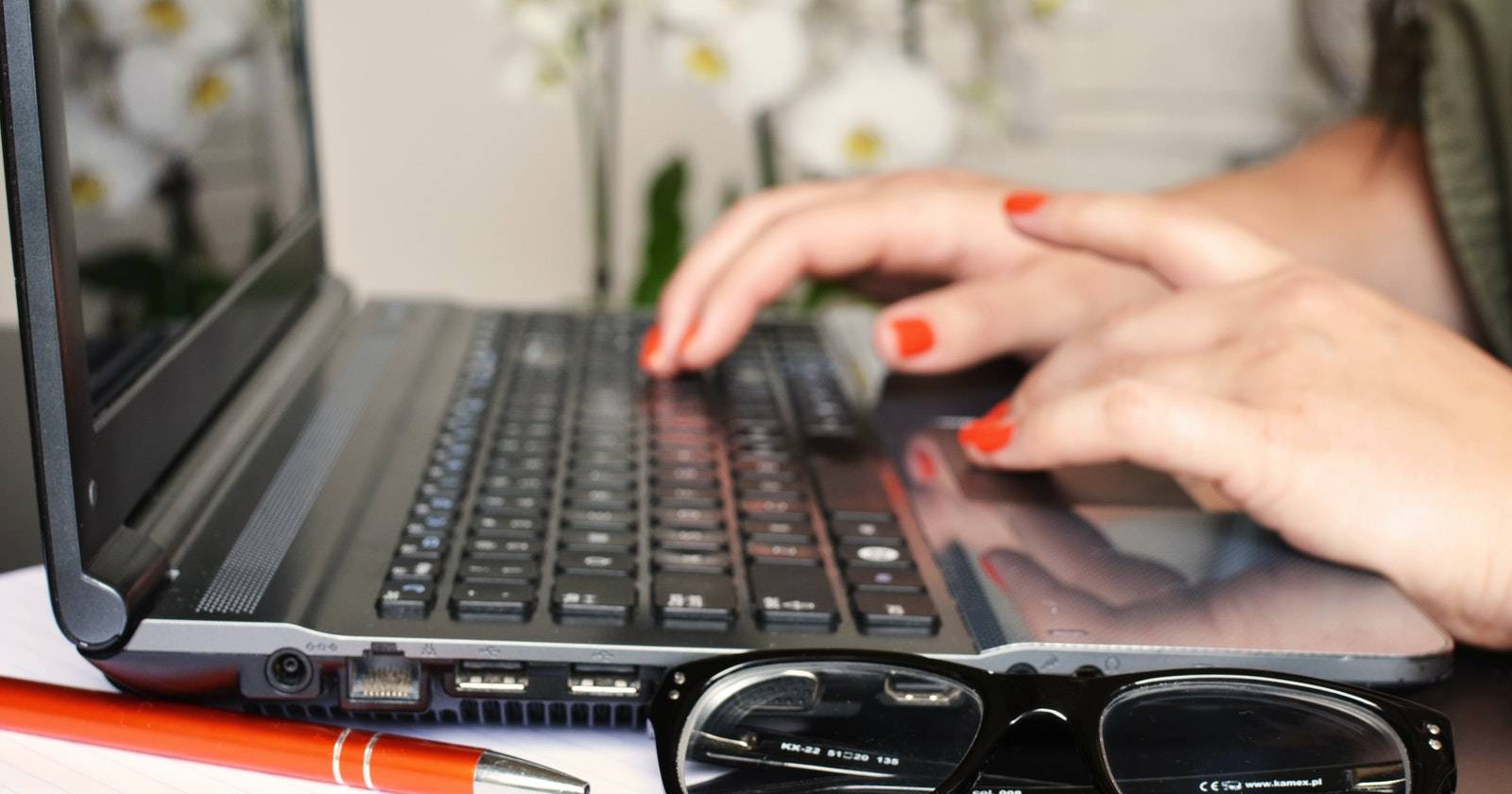 Женщина за ноутбуком с ручкой и очками на столе