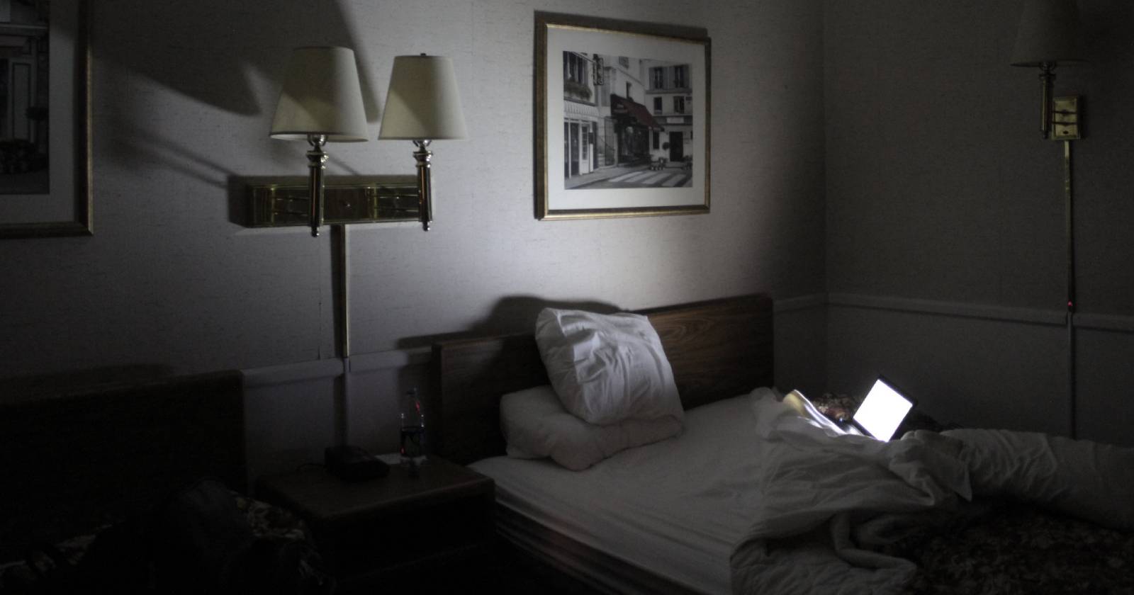 Открытый ноутбук на кровати освещает затемнённую гостиничную комнату.