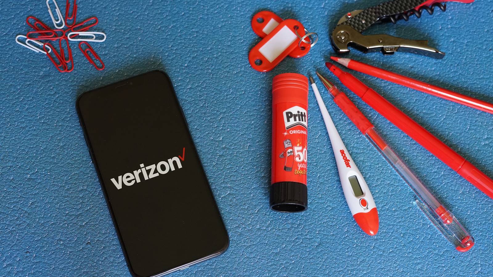 Логотип Verizon на экране iPhone рядом со скрепками и красными канцелярскими принадлежностями