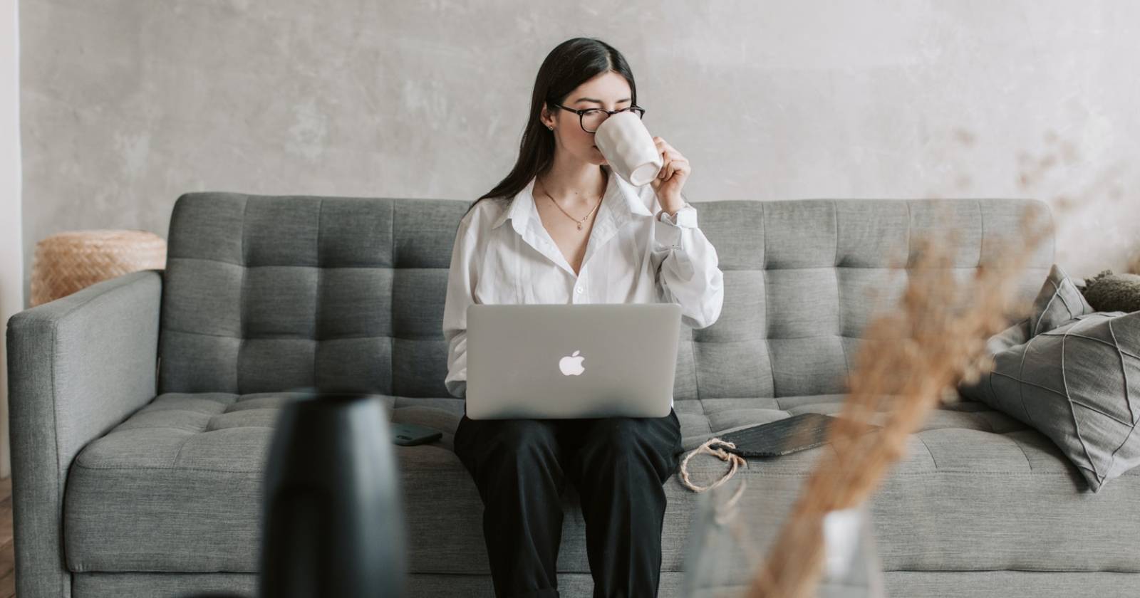 Женщина пьёт из белой кружки на диване с ноутбуком